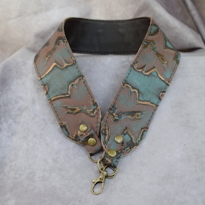 May include: A brown leather camera strap with a teal and gold pattern. The strap has two brass rivets and a brass swivel hook.