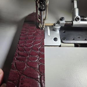 Puede incluir: Primer plano de una máquina de coser cosiendo una tira de cuero burdeos. El cuero tiene un patrón texturizado y en relieve. La aguja y los componentes metálicos de la máquina son visibles, destacando la costura y el detalle del material.