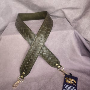以下が含まれることがあります： 金色の金属製の留め具が付いた、濃い緑色の革製の織りストラップ。ストラップはバッグや財布用です。ストラップは手作りで、アメリカ製です。タグには「Alford Designs」と「Handcrafted fine leather purses, bags, men's satchels, replacement straps, briefcases, accessories, and gifts. Made in the USA.」の文字が書かれています。
