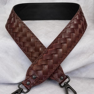 Op de afbeelding: Een brede tasriem met een buitenkant van donkerbruin gevlochten leer en een binnenkant van glad zwart leer. De riem is voorzien van twee matzwarte metalen draaibare karabijnsluitingen die met bijpassende klinknagels aan het gevlochten deel zijn bevestigd.
