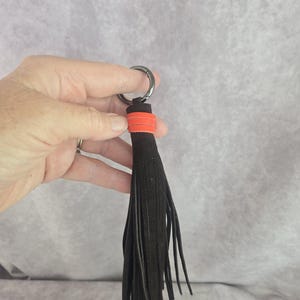 以下が含まれることがあります： 黒い革のタッセルキーホルダー。オレンジ色のアクセントとシルバー色のリングが付いています。タッセルは細長い革のストリップでできています。キーホルダーは、明るい灰色の背景に対して保持されています。