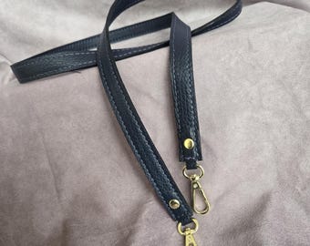 May include: A navy blue leather bag strap with gold-colored metal clasps. The strap has a textured surface and white stitching along the edges. The clasps are attached with small, gold-colored rivets.
