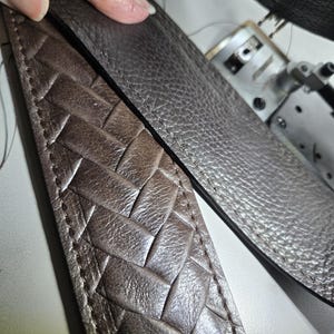 May include: A dark brown leather strap featuring a woven herringbone pattern on one side and a pebbled texture on the other. The strap is constructed with visible stitching along the edges, showcasing the contrast between the braided and smooth leather.