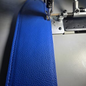 Op de afbeelding: Close-up van een naaimachine die een stuk felblauw leer naait. Het leer heeft een getextureerd oppervlak en wordt genaaid met een zilveren naald en draad. De focus ligt op het gedetailleerde vakmanschap van het naaiproces.