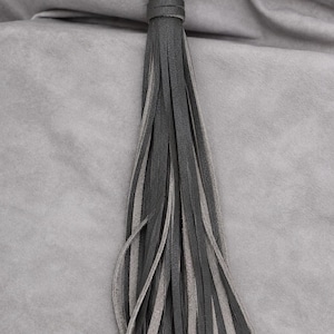 May include: A decorative tassel made of multiple thin strips of dark charcoal grey and light grey leather. The strips are gathered at the top with a matching leather band and attached to a circular antique brass-coloured metal ring clasp.