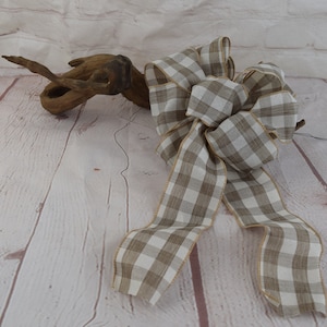 May include: A large, brown and white gingham bow with a gold wired edge. The bow is tied with a wire and is lying on a whitewashed wood surface.