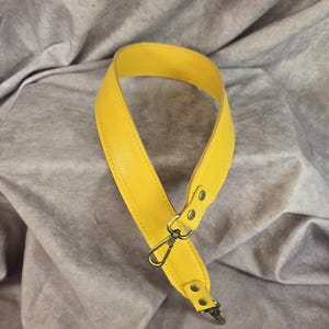 Puede incluir: Correa de bolso de cuero amarillo brillante con herrajes dorados. La correa presenta una superficie texturizada y está diseñada para sujetarse a un bolso. La correa tiene un cierre giratorio y mide aproximadamente 2,5 cm de ancho.