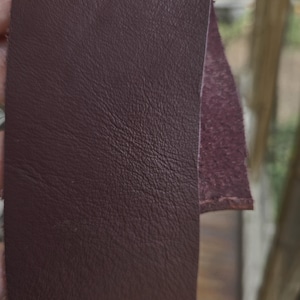 May include: A strip of dark burgundy leather with a smooth, slightly grained surface on the front side and a rough, fibrous texture on the exposed back edge. The material is flexible and shows a consistent deep red-brown color throughout.