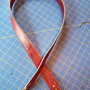 May include: A long, burgundy leather strap with gold metal hardware and a swivel clasp. The strap is lying on a blue gridded surface.