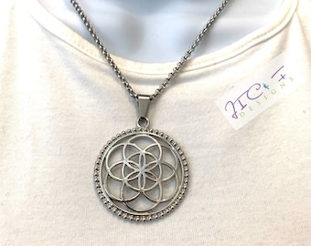 May include: A silver necklace with a round pendant featuring a geometric flower of life design. The pendant is surrounded by a beaded border.