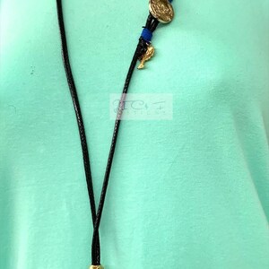 May include: A black cord necklace featuring a gold coin pendant, blue details, and a teal glass leaf-shaped charm. The necklace includes a gold bead and a small gold charm. The background is a light green top.