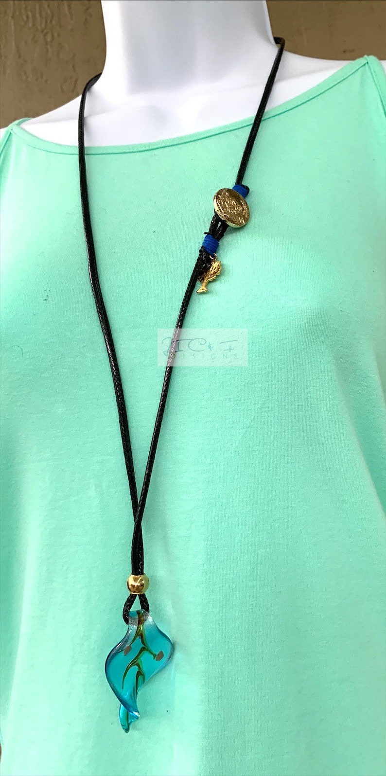 May include: A black cord necklace featuring a gold coin pendant, blue details, and a teal glass leaf-shaped charm. The necklace includes a gold bead and a small gold charm. The background is a light green top.