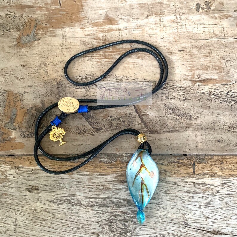 May include: A handmade necklace with a blue glass pendant with floral details. The pendant is suspended from a black cord, with gold and blue embellishments, including a gold tree charm and a circular gold clasp. The necklace is displayed on a wooden surface.