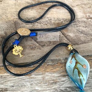 May include: A black cord necklace with a gold-coloured tree charm and a blue and gold accent. The necklace features a blue and clear glass pendant with brown and gold accents. The pendant is leaf-shaped.