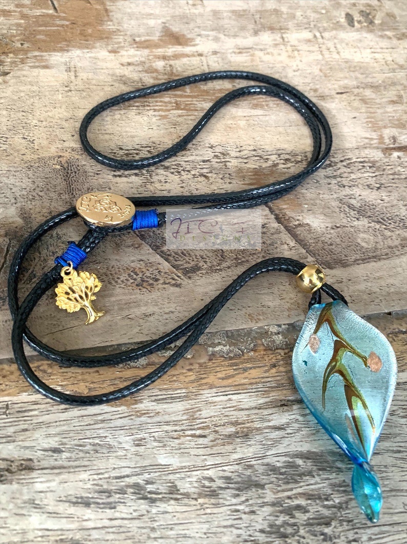 May include: A black cord necklace with a gold-coloured tree charm and a blue and gold accent. The necklace features a blue and clear glass pendant with brown and gold accents. The pendant is leaf-shaped.