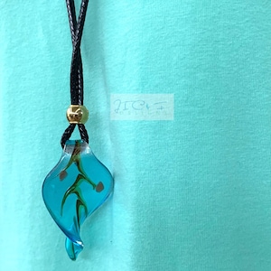 May include: A handmade necklace with a teal glass pendant, decorated with brown and green floral details. The pendant is suspended from a black cord with a gold-coloured bead. The image includes the text "JIC Designs".