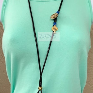 May include: A long black cord necklace with a teal glass pendant. The necklace features gold-coloured accents and a gold-coloured tree charm. A gold-coloured medallion and blue cord accents are also present.