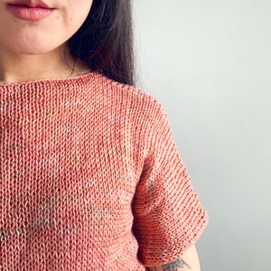 K&ouml;nnte beinhalten: Nahaufnahme einer Frau, die ein korallenfarbenes Strickoberteil mit kurzen &Auml;rmeln tr&auml;gt. Das Oberteil hat ein dezentes, gesprenkeltes Muster.