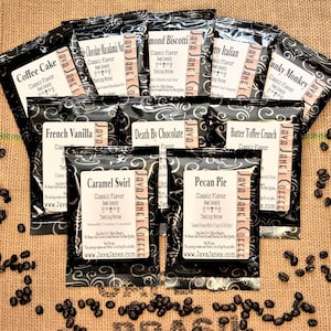 May include: An assortment of flavored coffee packets, including Coffee Cake, French Vanilla, and Pecan Pie, arranged on a burlap surface. Each packet is black with a white label, and coffee beans are scattered around the packets.