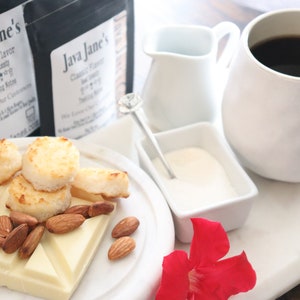 May include: A white mug filled with dark coffee sits next to a plate of treats. The plate holds toasted coconut macaroons, almonds, and a white chocolate bar. Two bags of coffee are in the background, along with a small creamer and sugar.