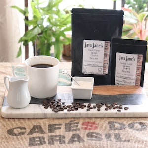 May include: A still life arrangement featuring coffee and coffee-related items. A white mug filled with coffee, a creamer, and a small dish of sugar sit on a marble serving board with coffee beans. Two black bags of coffee are labeled "Java Jane's".