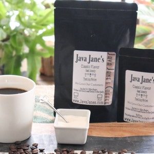 May include: A white mug filled with dark coffee sits next to a small white creamer and a square dish of sugar. Two black bags of coffee with the label "Java Jane's" are in the background, along with scattered coffee beans.