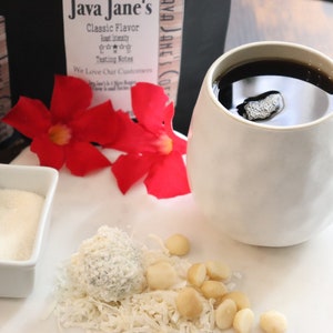 May include: A white ceramic mug filled with dark coffee, accompanied by a small dish of sugar, a spoon, a coconut ball, macadamia nuts, and red flowers. The background includes coffee packaging with the text "Java Jane's Classic Flavor".