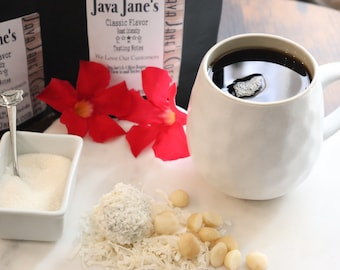 May include: A white ceramic mug filled with dark coffee, accompanied by a small dish of sugar, a spoon, a coconut ball, macadamia nuts, and red flowers. The background includes coffee packaging with the text "Java Jane's Classic Flavor".