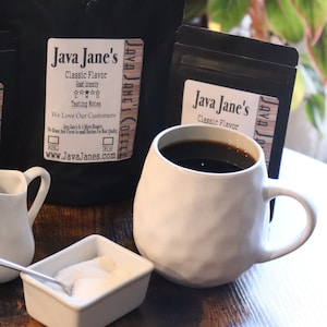 May include: A white ceramic mug filled with dark coffee sits next to a small dish of sugar and a creamer. Two black bags of coffee beans with the text "Java Jane's Classic Flavor" are in the background. The coffee is ready to be enjoyed.