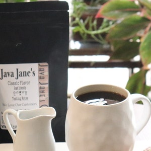 May include: An image of coffee-related products. Two black bags of coffee with the text "Java Jane's Classic Flavor" and a white creamer pitcher are in front of a white mug filled with coffee. The background includes a plant.