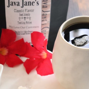 May include: A white mug filled with dark coffee, with a small foam bubble on top. The image includes a small dish of sugar, a decorative spoon, red flowers, and a package of Java Jane's coffee.