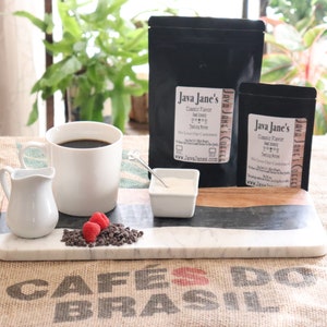 May include: A coffee setup featuring a white mug filled with dark coffee, a small creamer, and a serving of chocolate chips and raspberries. Two black bags of coffee beans with the text "Java Jane's" are also present, along with a small dish of cream.