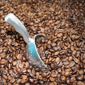 May include: A close-up shot of roasted coffee beans filling the frame. A silver scoop rests among the beans, partially filled. The beans are a rich brown color, with variations in tone and texture. The image suggests a coffee shop or a bulk food setting.