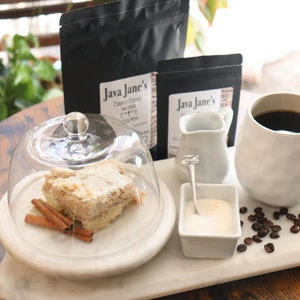 May include: A coffee setup featuring a white mug filled with dark coffee, a small creamer, sugar, and coffee beans. A slice of coffee cake is under a glass dome, with cinnamon sticks. Two bags of coffee beans are in the background.