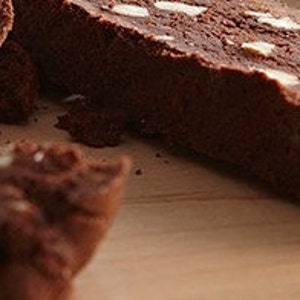 May include: Close-up of several slices of chocolate biscotti with visible almond pieces. The biscotti are a rich brown color and sit on a light brown wooden surface. The image highlights the texture and ingredients of the baked goods.
