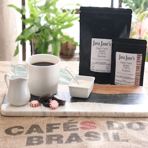 May include: A coffee setup featuring a white mug filled with dark coffee, a small creamer, and a marble serving board. The board holds cookies, mints, and a small bowl of sugar. Two black bags of coffee beans with the text "Java Jane's" are also present.