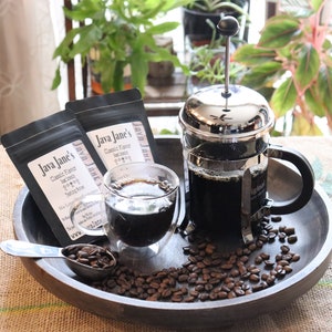 May include: A coffee setup featuring a French press, a glass of coffee, and two bags of Java Jane's coffee. Coffee beans are scattered around a wooden tray with a measuring spoon. The scene is set on a burlap surface.