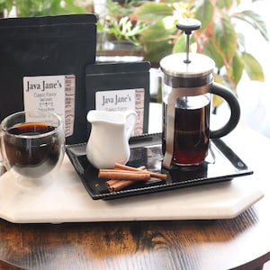May include: A coffee setup featuring a French press, a double-walled glass mug with coffee, a small creamer, and cinnamon sticks on a tray. Two black coffee bags with the text "Java Jane's" are also present. The setup is on a white marble tray.