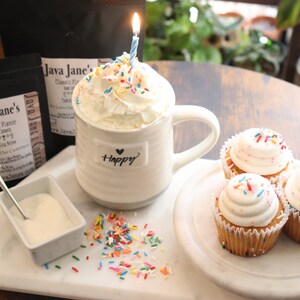 May include: A festive scene featuring a white mug with whipped cream, sprinkles, and a lit candle. The mug has the word "happy" on it. There are cupcakes with sprinkles and bags of coffee beans. A small dish of sugar and a spoon are also present.