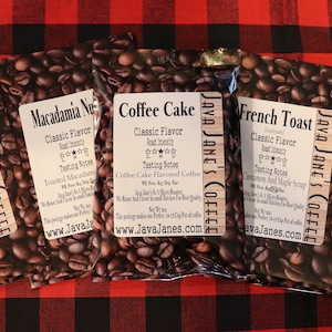 May include: Five coffee bean bags with labels, each featuring a different flavor: Vanilla Hazelnut, Macadamia Nut, Coffee Cake, French Toast, and Butterscotch Toffee. The bags are on a red and black plaid surface.