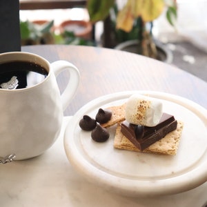 May include: A white mug filled with dark coffee sits next to a plate with a s'more and chocolate candies. A small container of sugar and a spoon are also on the table. A bag of coffee beans is in the background.