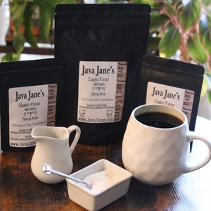 May include: An arrangement of coffee-related items on a wooden table. Includes black coffee in a white mug, small creamer, sugar bowl with a spoon, and several black bags of coffee with the text "Java Jane's Classic Flavor".