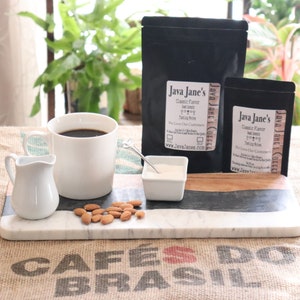May include: A coffee setup featuring a white mug filled with coffee, a small creamer, and a bowl of almonds. Two black bags of coffee beans with the text "Java Jane's" are also present. The setup is on a marble and wood serving board.