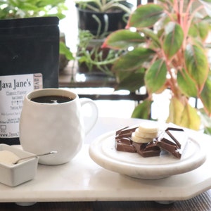 May include: A white mug filled with dark coffee, a small dish of sugar, and a plate of chocolate and banana slices sit on a white tray. A black bag of Java Jane's coffee is in the background. Green plants are visible.