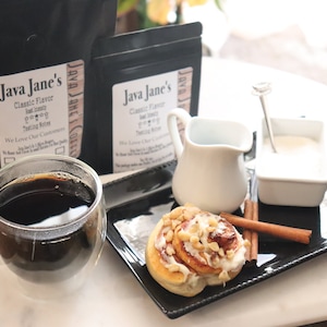 May include: A clear glass mug filled with dark coffee sits next to a black tray holding a cinnamon roll, cream, and sugar. Two bags of coffee beans with the text "Java Jane's" are in the background. A cozy coffee shop scene.