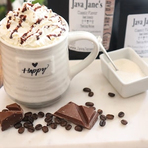 May include: A white ceramic mug filled with whipped cream and chocolate syrup, with the word "Happy" and a heart on the front. Coffee beans and chocolate pieces are scattered around the mug. Java Jane's coffee bags are in the background.