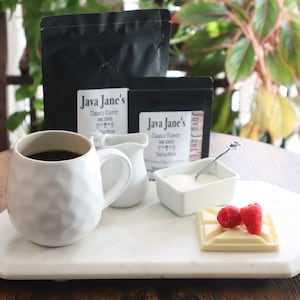 May include: A white ceramic mug filled with dark coffee sits on a marble tray, alongside a small creamer, sugar bowl, and a white chocolate square topped with raspberries. Two bags of coffee beans are in the background.