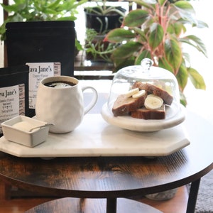 May include: A still-life image featuring a white mug filled with coffee, two black coffee bags, and a small dish of cream. Sliced banana bread is under a glass cloche on a marble tray. The text on the coffee bag reads "Java Jane's Classic Flavor".