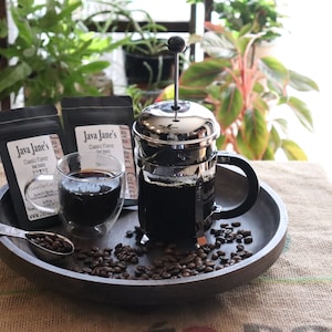 May include: A coffee setup featuring a French press, a double-walled glass of coffee, and two bags of Java Jane's Classic Flavor coffee. Coffee beans and a spoon are on a wooden tray, with plants in the background.