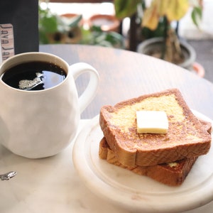 May include: A white mug filled with dark coffee sits next to a plate of French toast topped with butter. A bag of coffee beans and a small creamer are also visible. The coffee mug has a textured surface.
